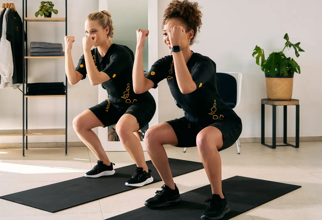 Schnelle Ergebnisse mit EMS-Training in Leipzig Zwei Frauen in EMS-Anzügen machen Kniebeugen und nutzen das EMS-Training in Leipzig, um in kurzer Zeit schnelle Resultate zu erzielen.